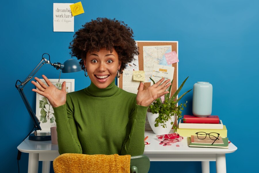 A black woman sits excitedly in front of a vision board filled with images, quotes, maps and goals. Her expression radiates enthusiasm and determination, reflecting her focus on achieving her dreams.