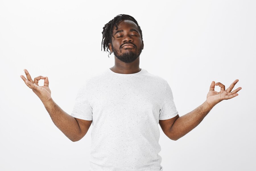 A serene black man wearing a white outfit smiles calmly in a minimalist white room. His eyes are closed, and his hands form a relaxation sign with his thumb and forefinger touching, exuding a peaceful and tranquil aura.