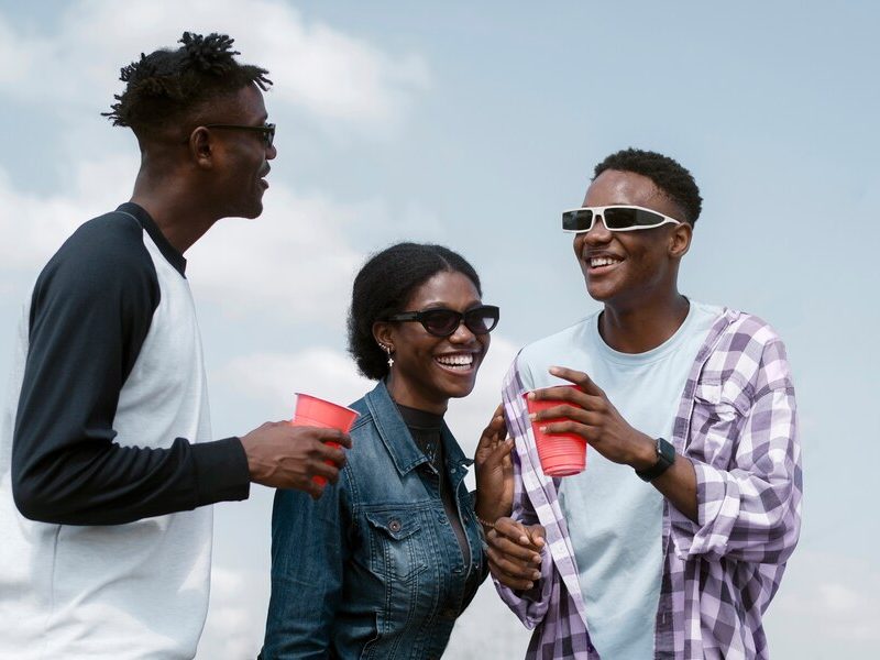 Two young black men and one black lady smile and laugh together, looking relaxed and having fun. They hold red cups, wearing sunshades, enjoying the moment in a carefree and joyful atmosphere.