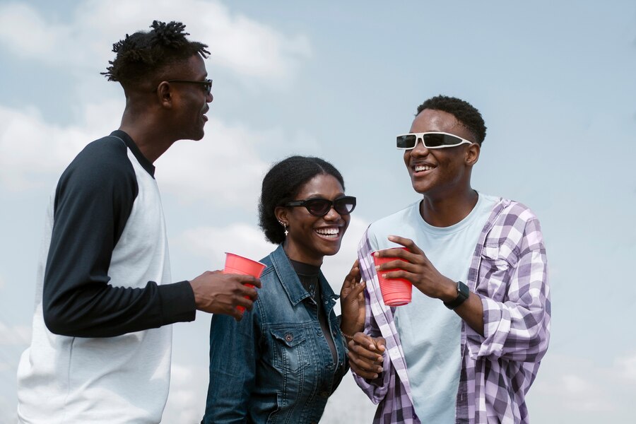 Two young black men and one black lady smile and laugh together, looking relaxed and having fun. They hold red cups, wearing sunshades, enjoying the moment in a carefree and joyful atmosphere.