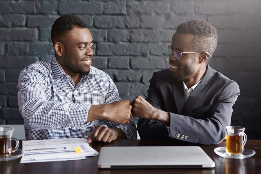 Two Black men smiling and exchanging a "knuckle chop" in celebration of a successful negotiation, with documents and papers spread out on the table in front of them.