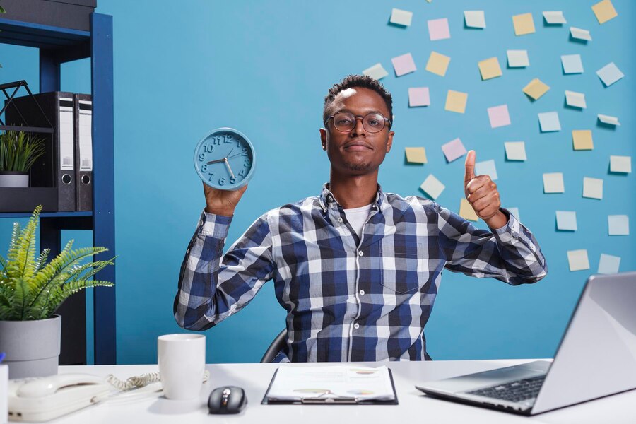 A confident Black man holding an alarm clock in one hand and giving a thumbs-up with the other, standing in front of a wall with sticky notes, symbolizing personal effectiveness and time management.