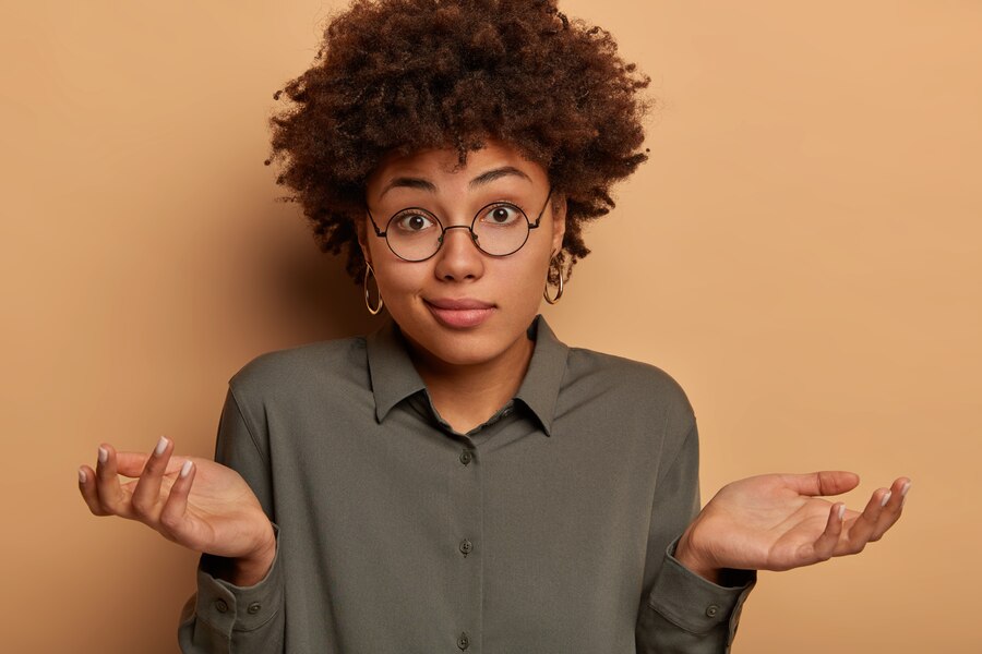 A Black woman with a curious expression shrugs her shoulders, her palms turned upward as if asking 'Why?' Her eyebrows are raised slightly, emphasizing her puzzled and questioning demeanor