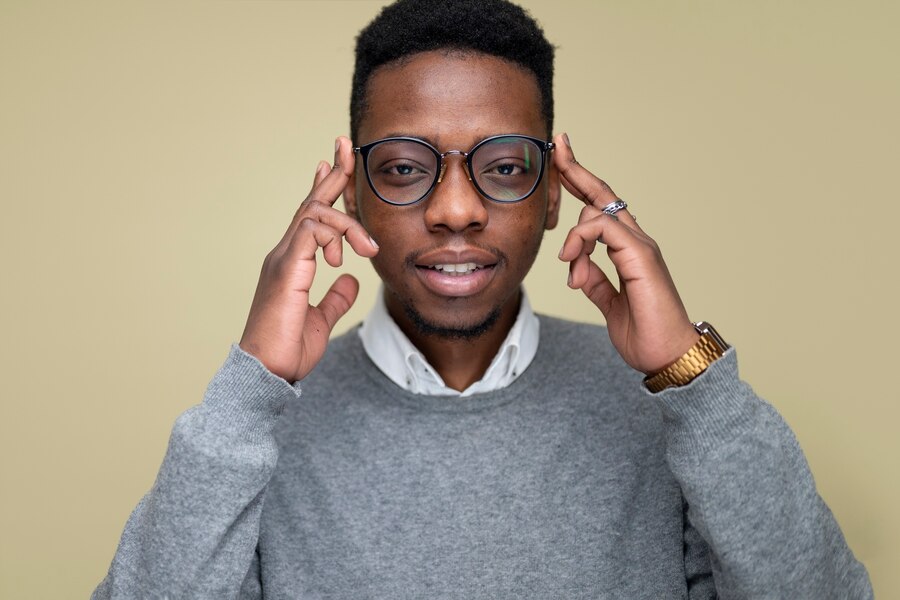 A front-facing Black man wearing glasses and smiling warmly, holding the sides of his head in a thoughtful gesture, conveying confidence and emotional intelligence.