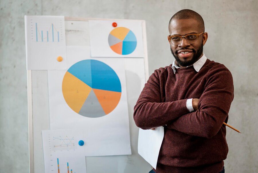 A confident Black man wearing glasses, folding his arms and delivering a presentation with poise and enthusiasm, standing in front of a board with pie charts.