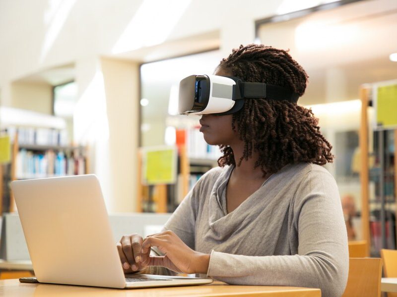 A young Black woman sitting in a library, wearing a virtual reality headset and typing on a laptop, surrounded by bookshelves and a warm, academic atmosphere.