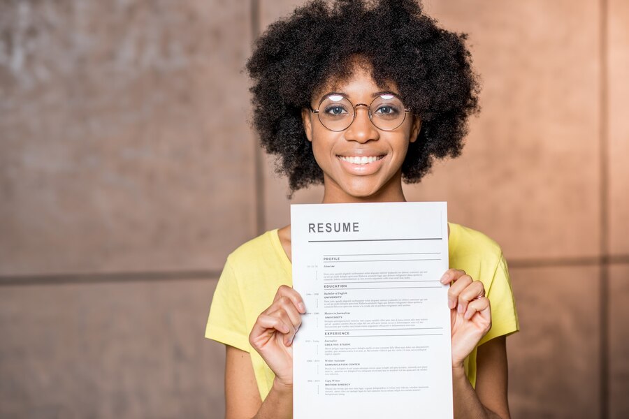 Young black female wearing with an afro, wearing yellow shirt and glasses holds her cv/resume while smiling.