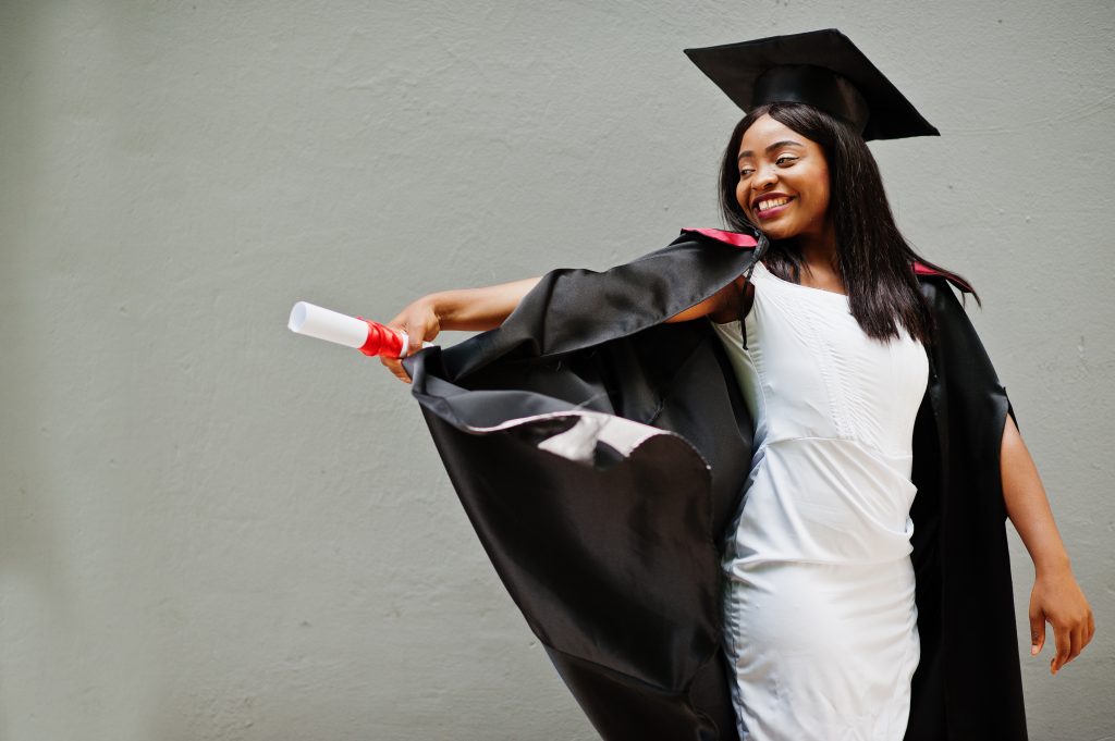 Young female african student wearing a graduation gown and hat with a scroll poses outdoors.